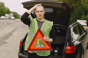 muž v reflexní vestě stojící u otevřeného kufru auta s výstražným trojúhelníkem v ruce